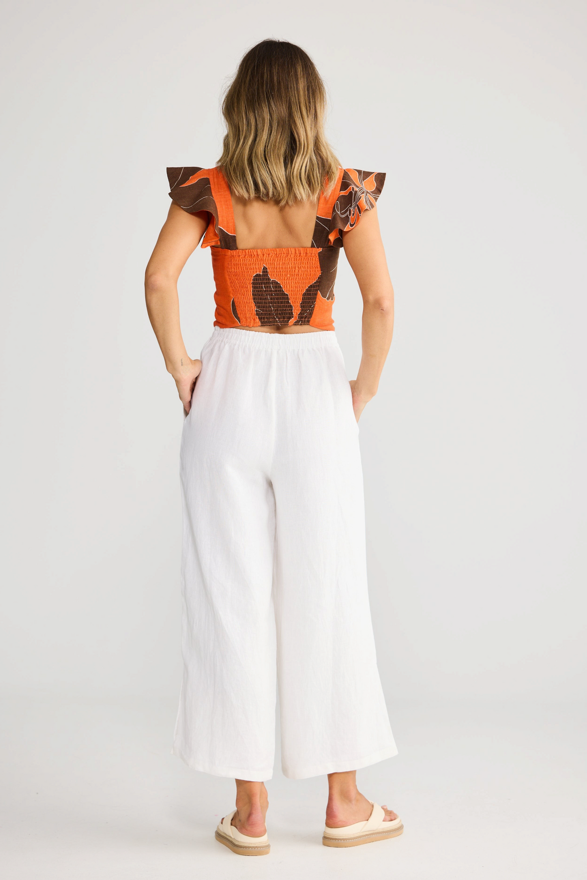 ELSHAM BOUTIQUE Zaria Top in Hibiscus Breeze orange floral linen with ruffle sleeves, worn with white linen pants.