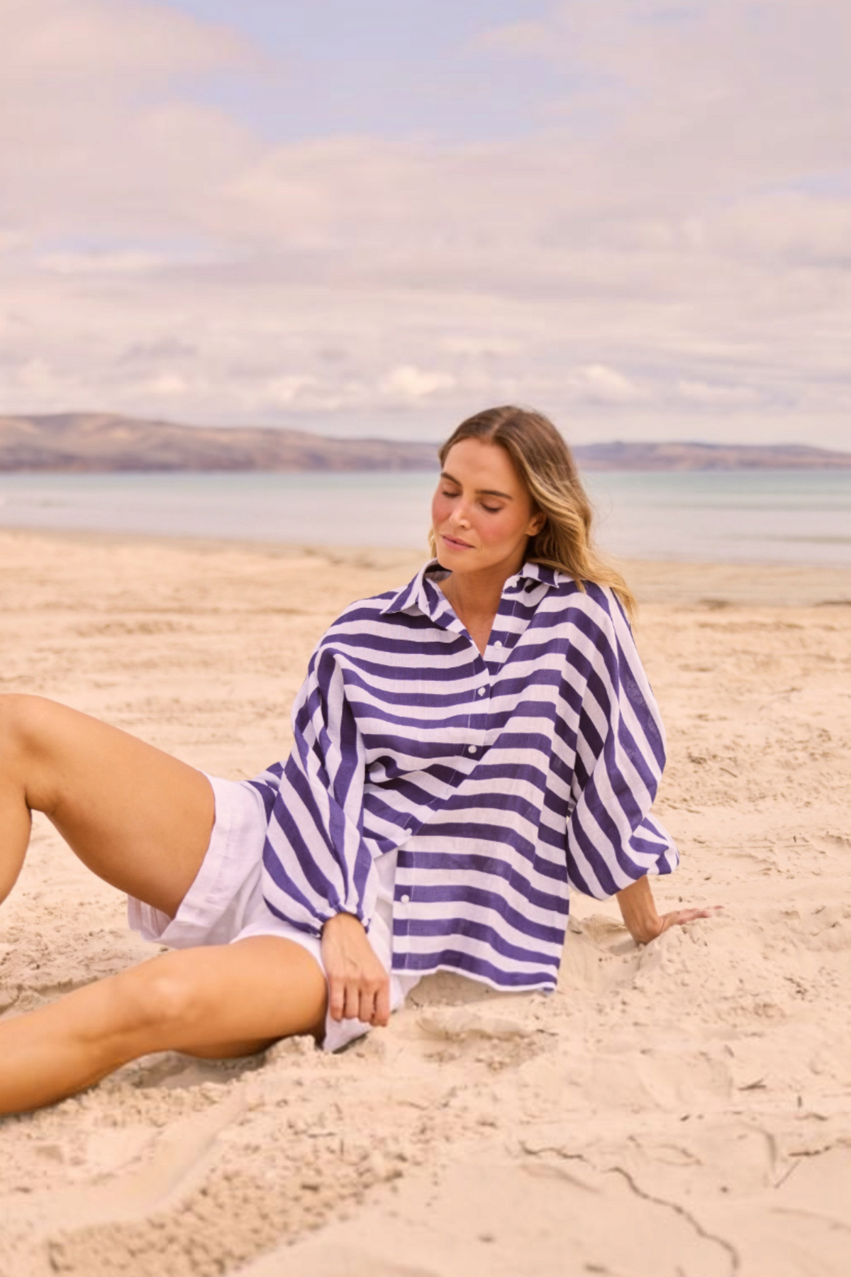 ELSHAM BOUTIQUE Cliffside Shirt in navy and white striped linen with oversized fit, worn by a woman on a sandy beach.