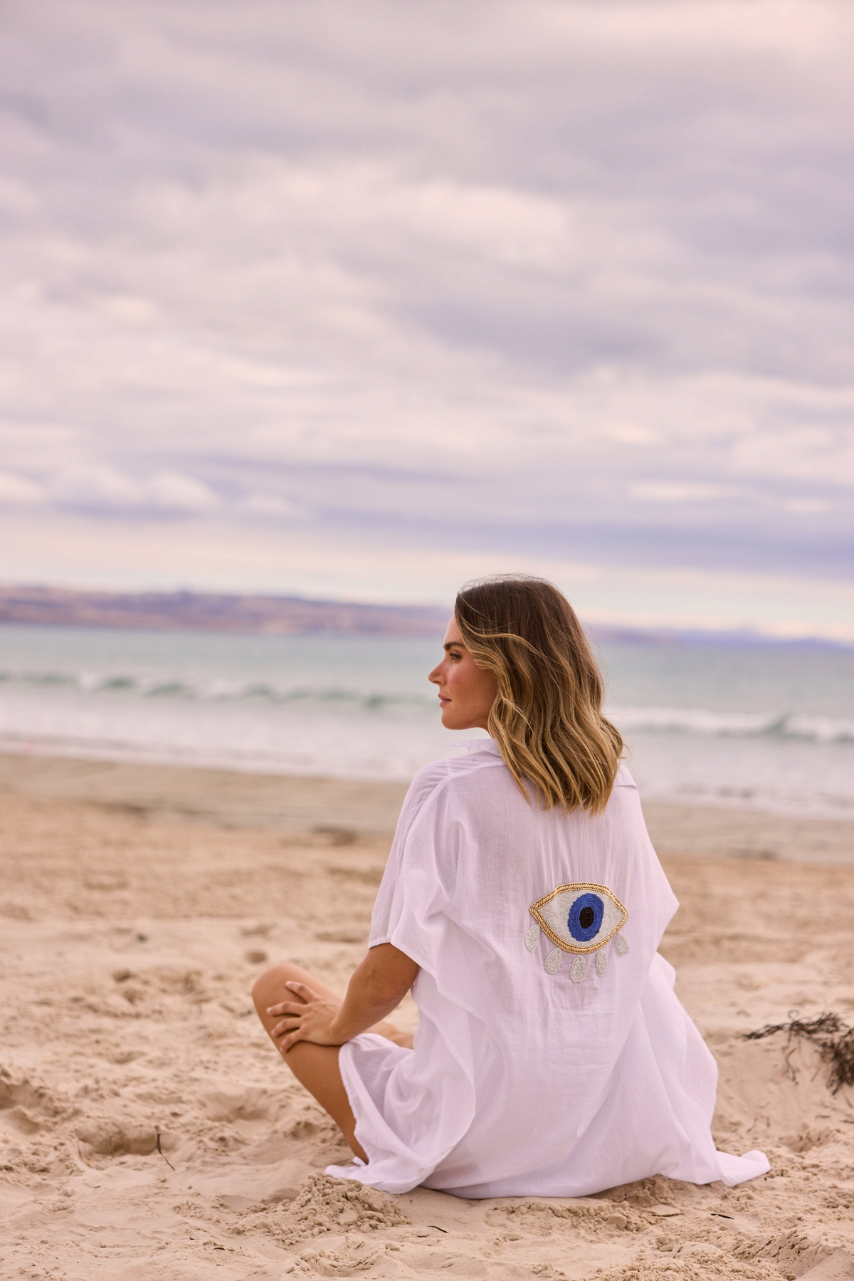 ELSHAM BOUTIQUE white cotton kaftan with evil eye embroidery on back, worn by woman sitting on a sandy beach.