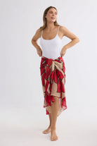 Model wearing an ELSHAM BOUTIQUE crimson floral cotton sarong over a white bodysuit against a white background