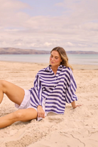ELSHAM BOUTIQUE Cliffside Shirt in navy and white striped linen with oversized fit, worn by a woman on a sandy beach.