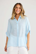 ELSHAM BOUTIQUE light blue oversized linen shirt with a collared neckline and short sleeves worn by a smiling woman.