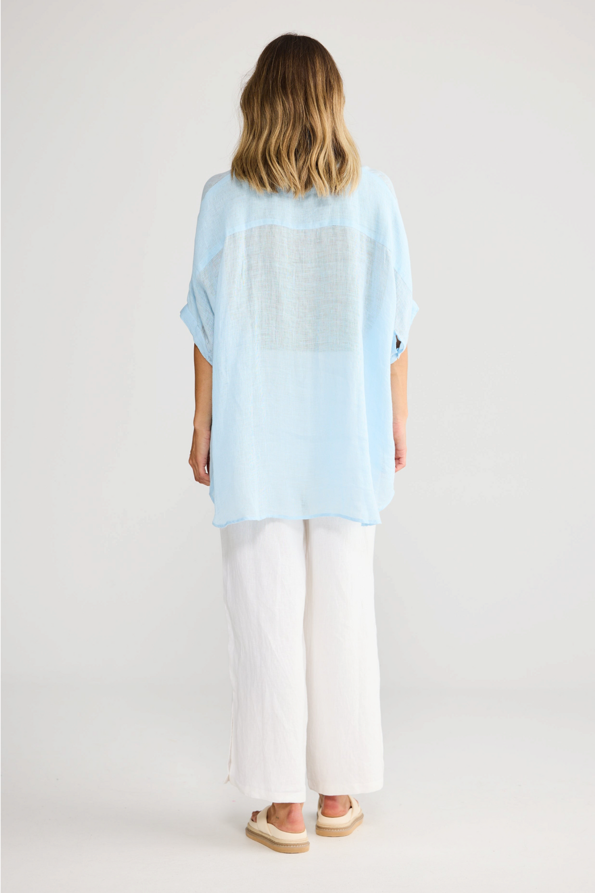 ELSHAM BOUTIQUE light blue oversized linen shirt with short sleeves, back view on a model wearing white trousers.