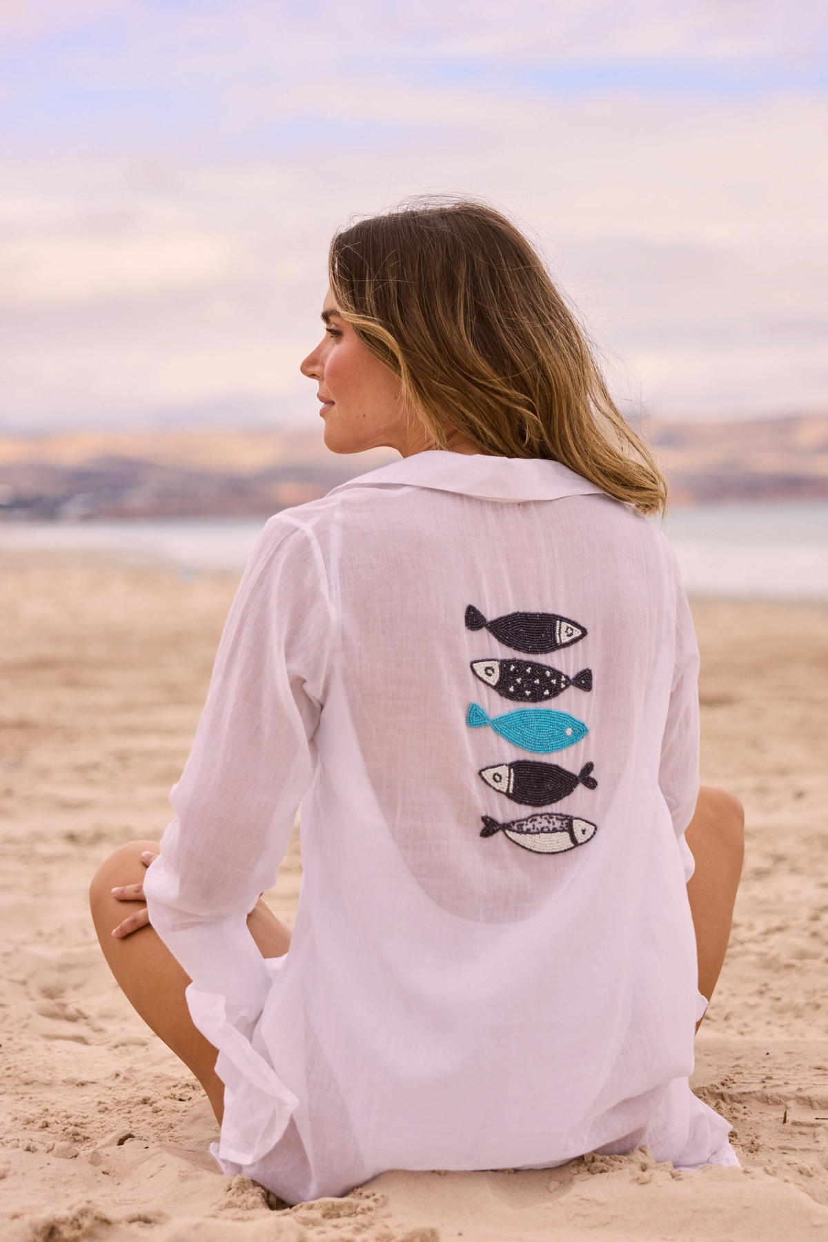 ELSHAM BOUTIQUE white cotton Lilo shirt dress with beaded fish embroidery on the back, worn by a woman on the beach.