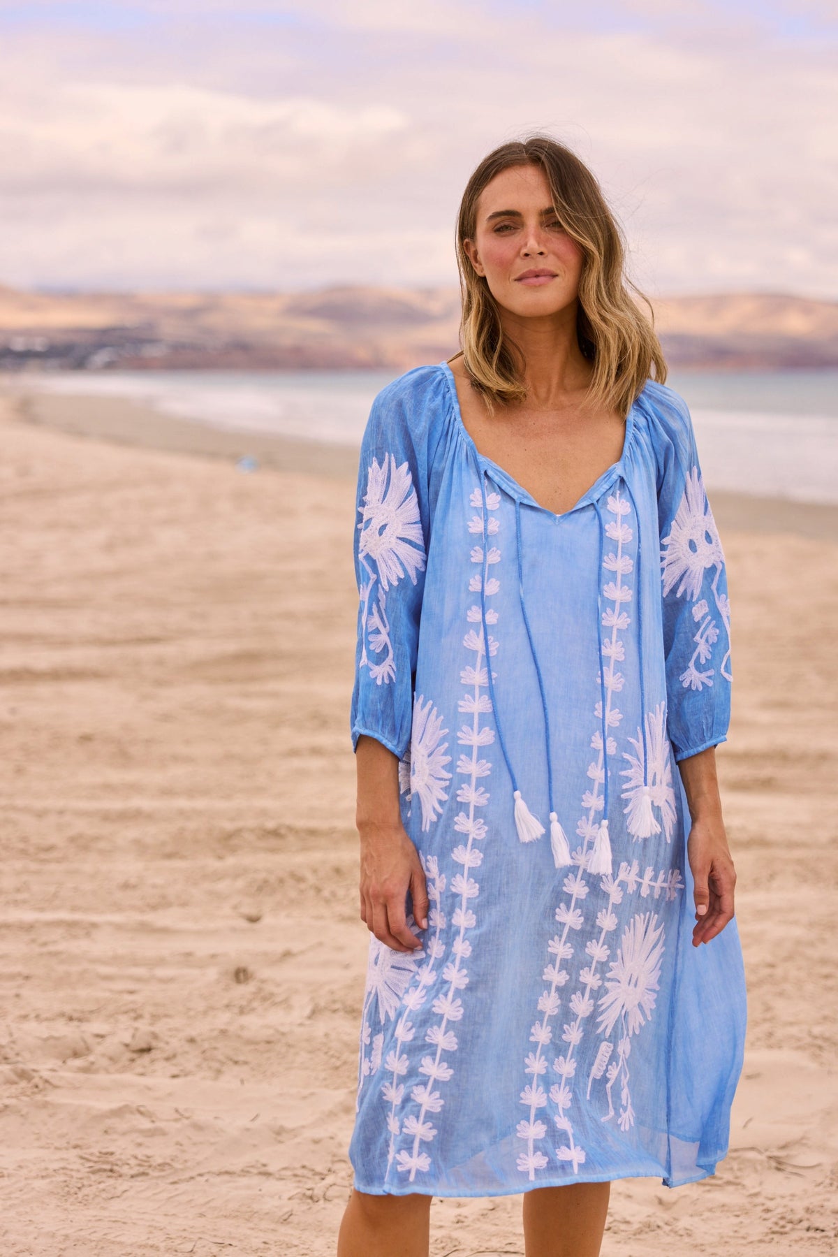 ELSHAM BOUTIQUE blue cotton knee-length Aloha dress with white floral embroidery and tie-front neckline on a beach.