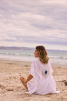 ELSHAM BOUTIQUE white cotton kaftan with evil eye embroidery on back, worn by woman sitting on a sandy beach.